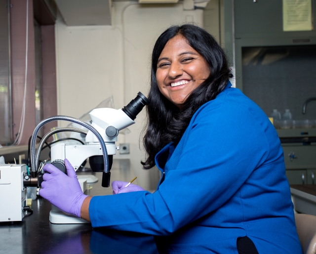 A smiling lab student using a microscope.